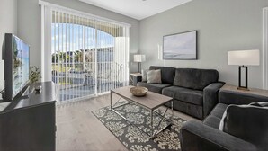 Una sala de estar moderna con un sofá gris, una mesita de centro y un televisor. Tiene un balcón con persianas blancas y vistas al exterior.