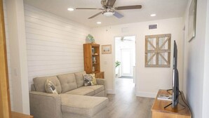 A living room with a sofa, a wooden cabinet, a ceiling fan, and a television on a wooden stand. - Cornerstone House (St. Augustine)