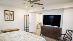A bedroom with a bed, a dresser, a TV, a ceiling fan, and a door leading to a bathroom. - Cornerstone House (St. Augustine)