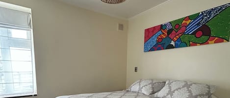 Un dormitorio con cama, ventana, una obra de arte en la pared y una lámpara en el techo.