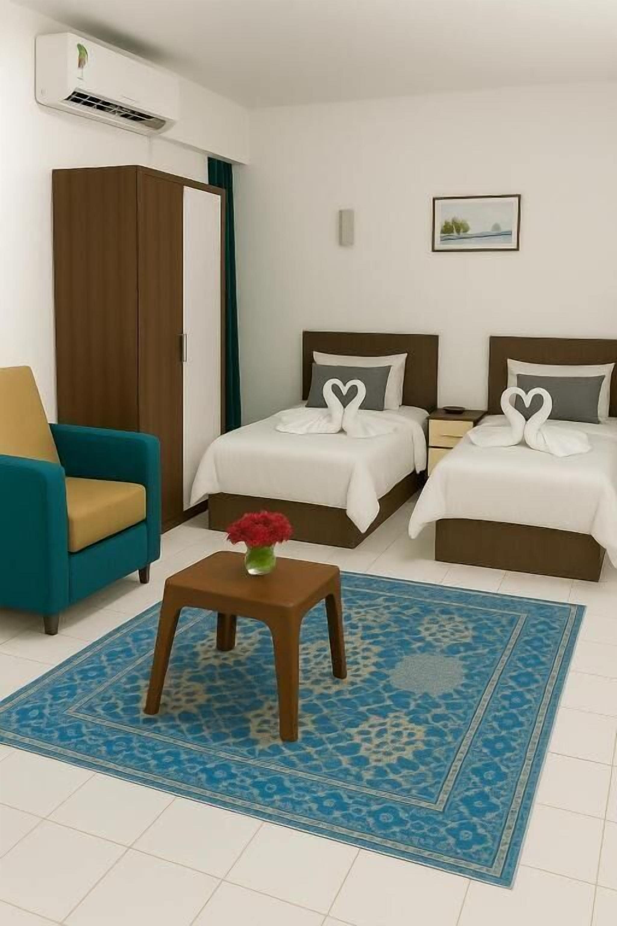 Une chambre d’hôtel avec deux lits, un tapis bleu à motifs, une petite table sur laquelle est posé un vase de fleurs, un fauteuil turquoise et une armoire.