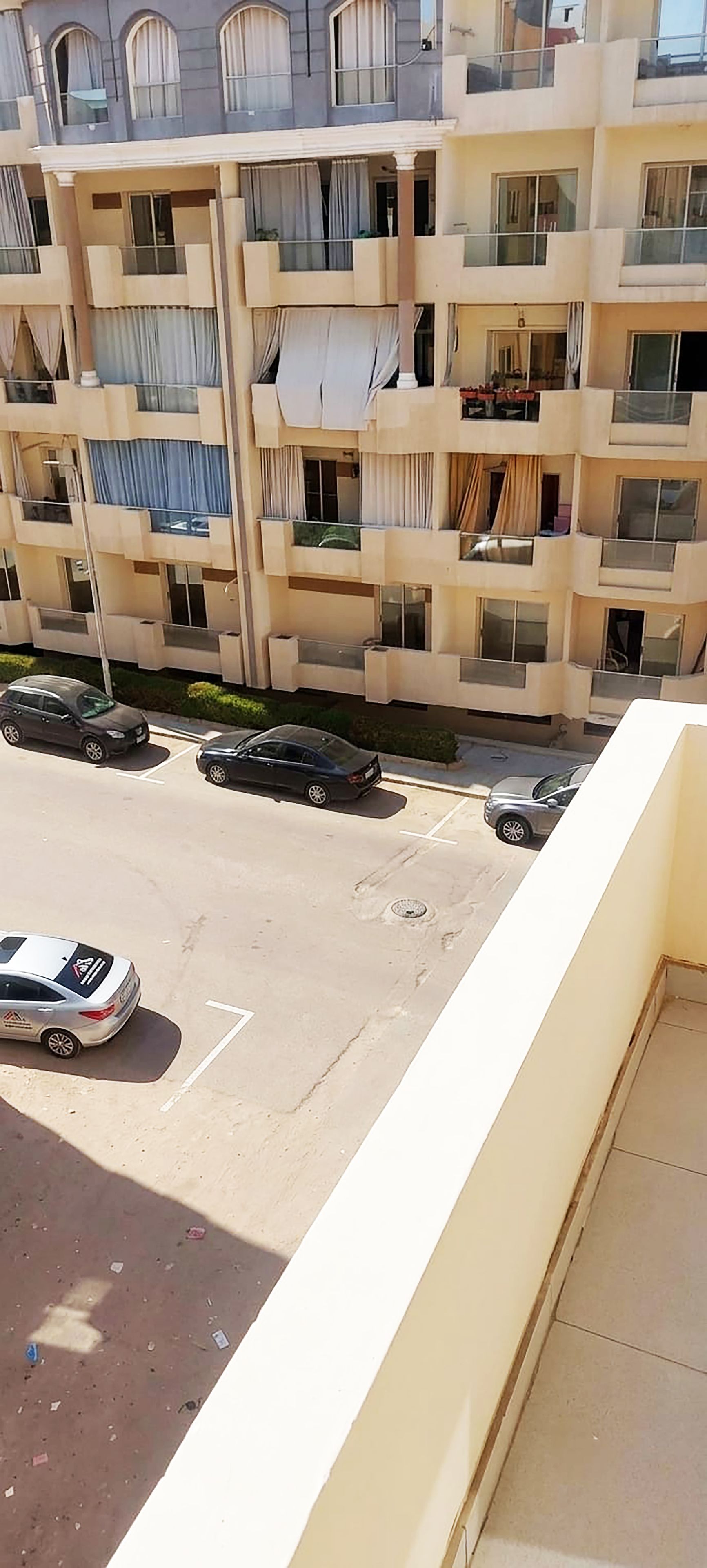 Un edificio residencial de varios pisos con balcones, un estacionamiento con coches y una vista despejada de la calle.