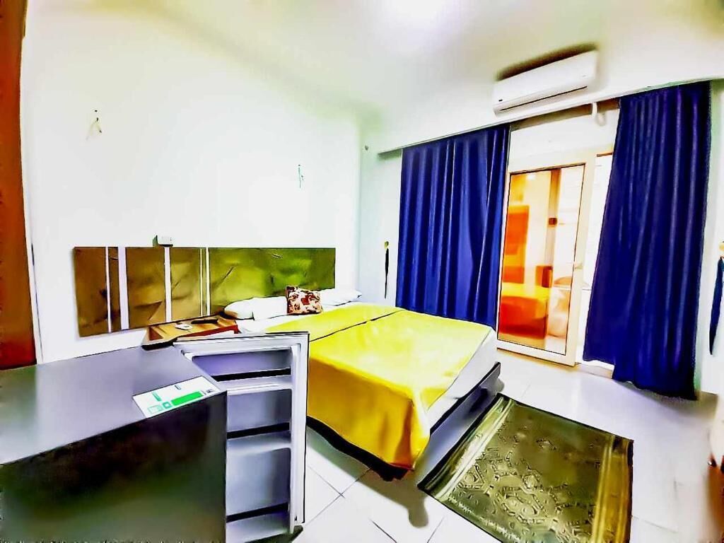 Quarto de hotel com cama, escrivaninha, cadeira, janela com cortinas e aparelho de ar condicionado.