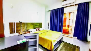 Quarto de hotel com cama, escrivaninha, cadeira, janela com cortinas e aparelho de ar condicionado.