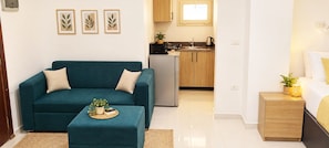 A living room with a teal sofa, a small ottoman, and a coffee table with a plant. There is a kitchen in the background with wooden cabinets and a sink. - Majestic apartments (Hurghada)