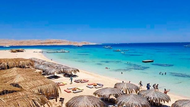Una playa con sombrillas de paja y tumbonas, agua turquesa cristalina y barcos a lo lejos.