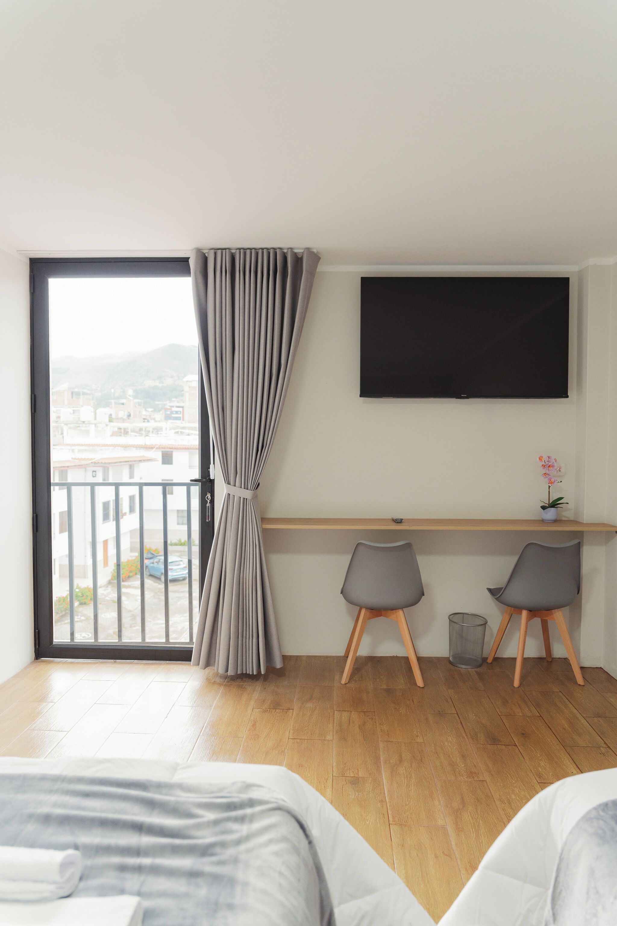 A modern hotel room with a large bed, a desk with two chairs, a TV, and a balcony with a view of buildings and mountains.