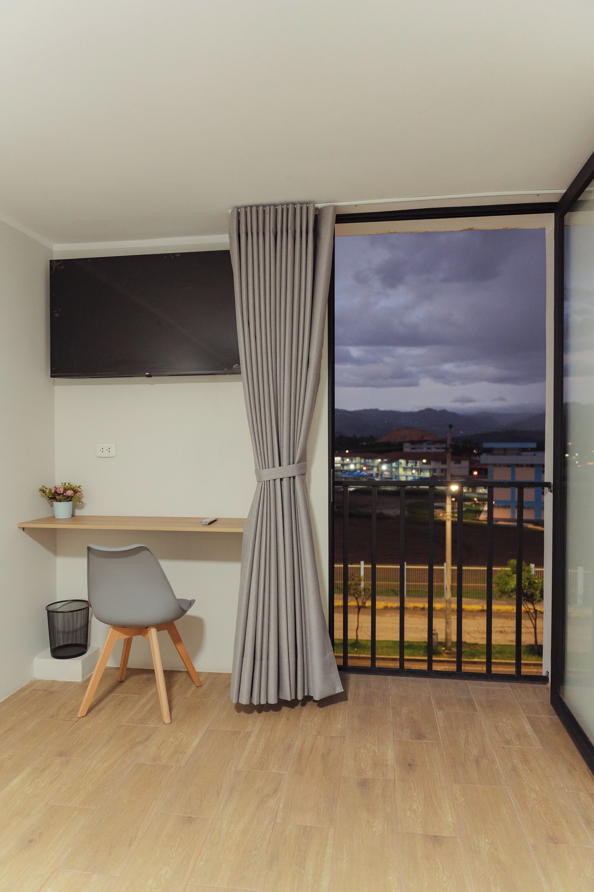 A modern room with a wooden desk, a grey chair, a mounted TV, and a balcony with a view of the city and mountains.