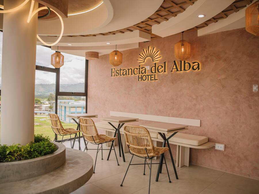 Un vestíbulo de hotel con sillas de mimbre, una mesa larga y un amplio ventanal con vistas a edificios y vegetación.