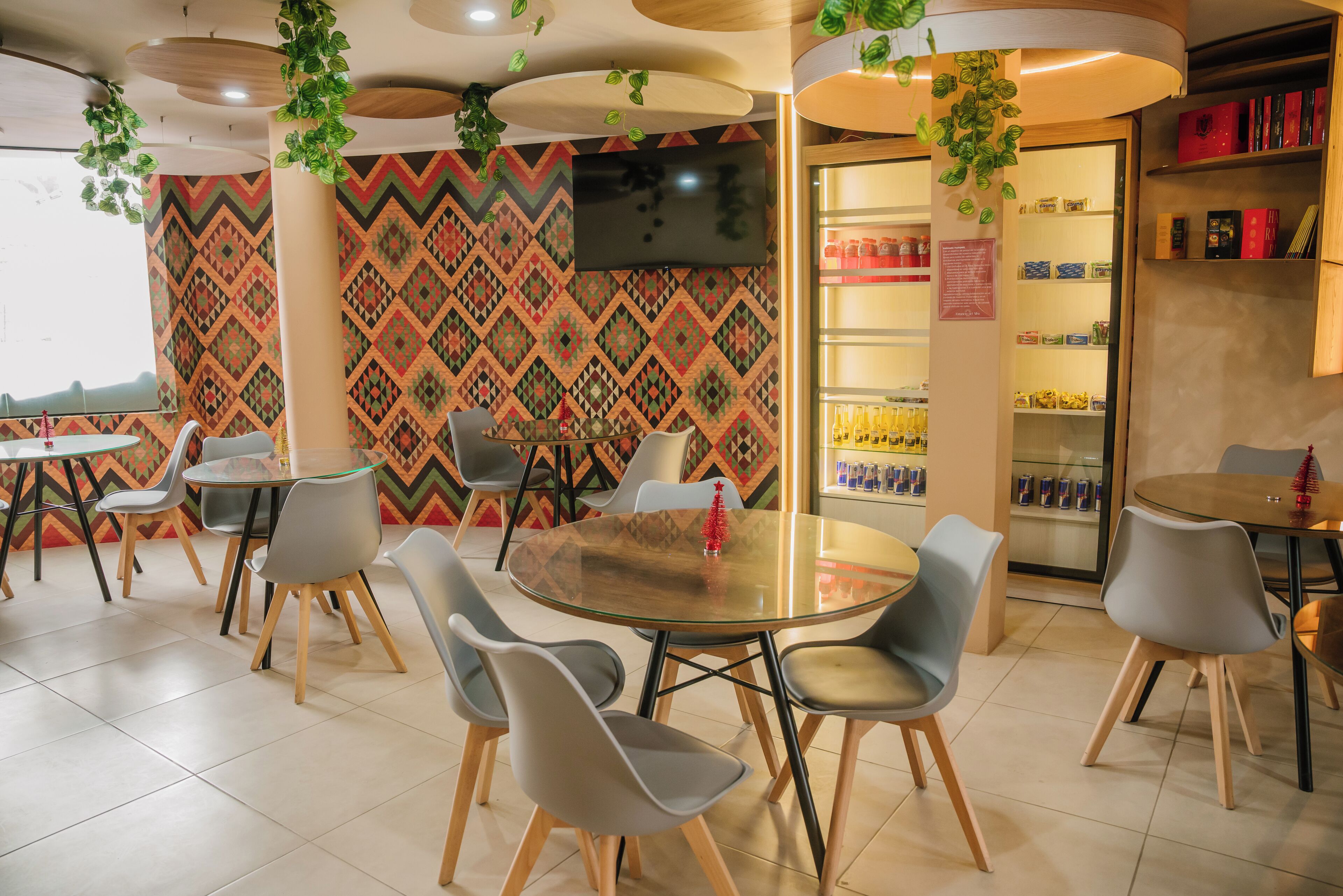 A modern cafe with round tables and grey chairs, a patterned wall, a TV, and a refrigerator with beverages.