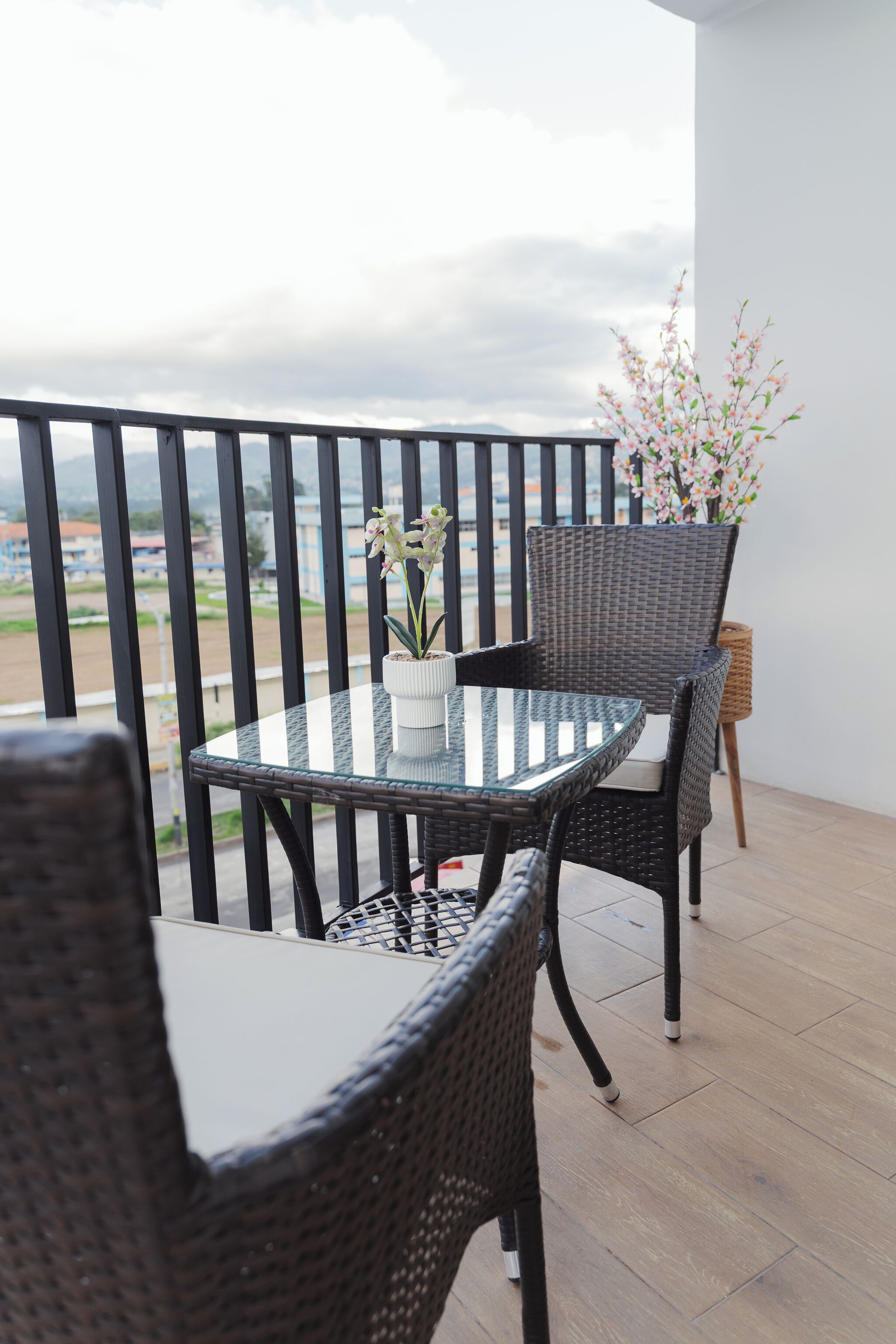 A balcony with a glass table and wicker chairs, overlooking a view of buildings and a distant landscape.
