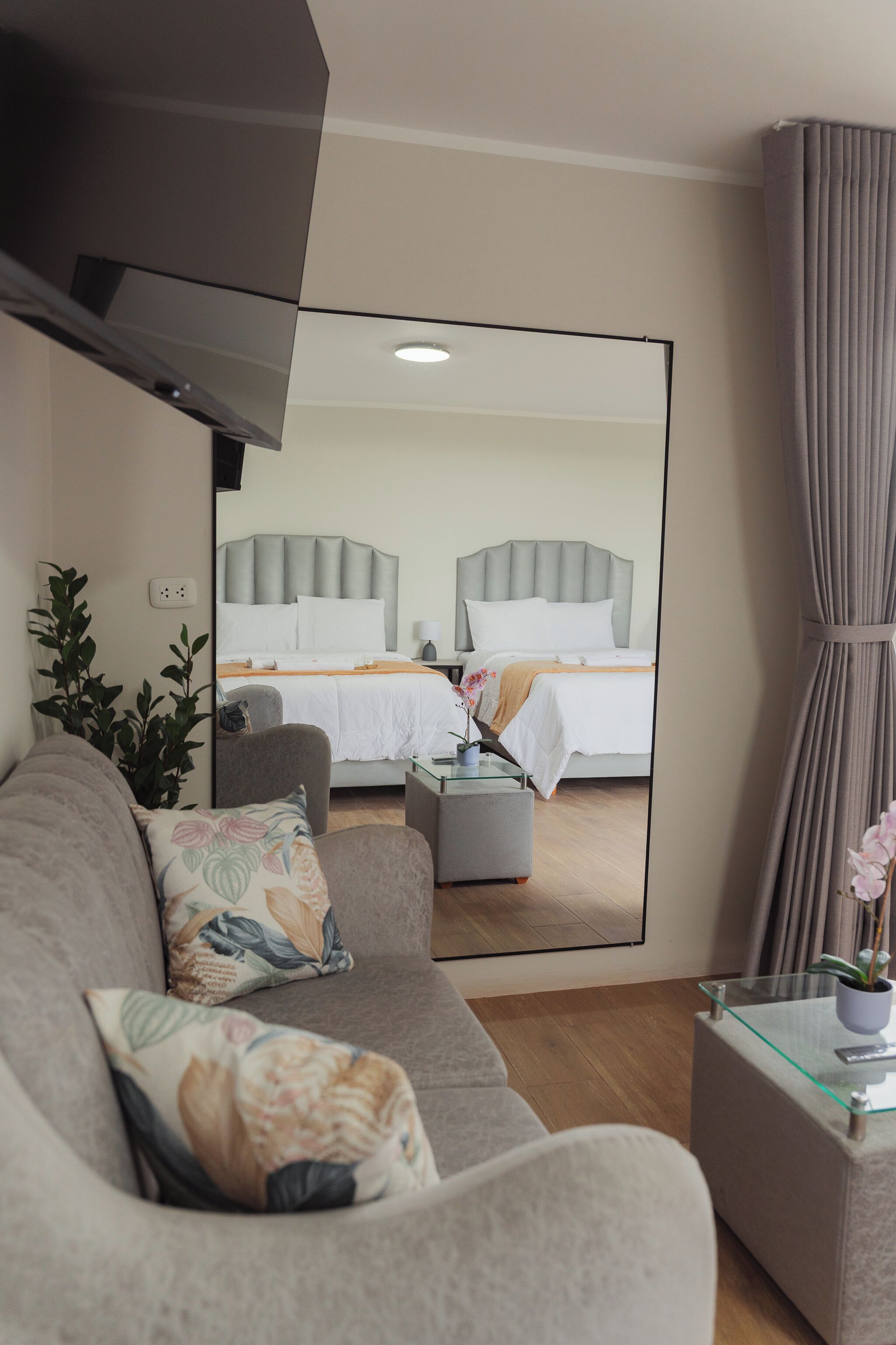 A modern hotel room with a large mirror reflecting a neatly made bed, a grey sofa, and a glass coffee table with a plant.