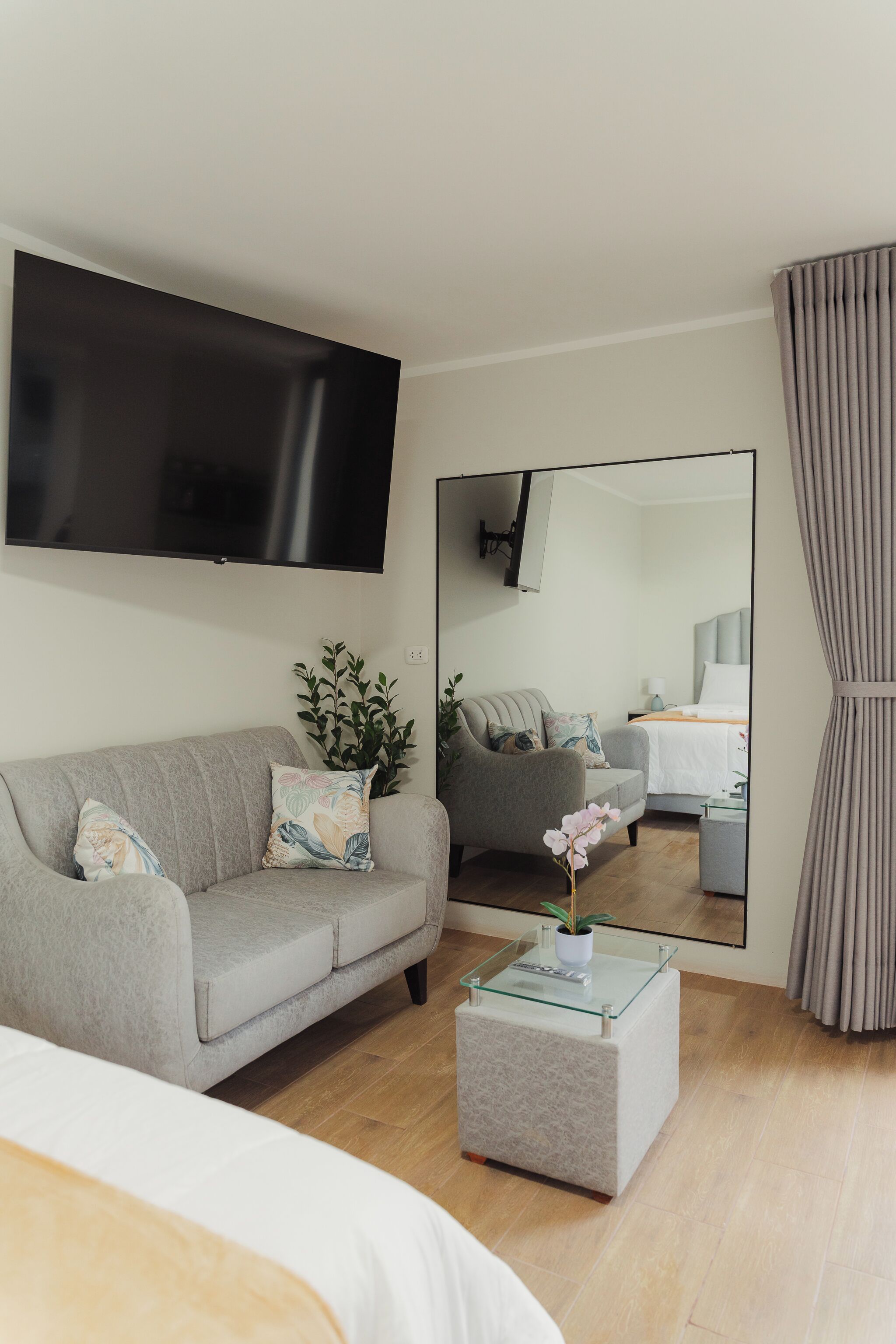 A modern hotel room with a large flat-screen TV mounted on the wall, a grey sofa with decorative pillows, a small glass coffee table with a potted plant, and a large mirror reflecting the room.