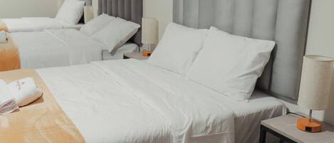 Habitación de hotel con dos camas, cada una con cabecero gris, ropa de cama blanca y una mesita de noche con lámpara.