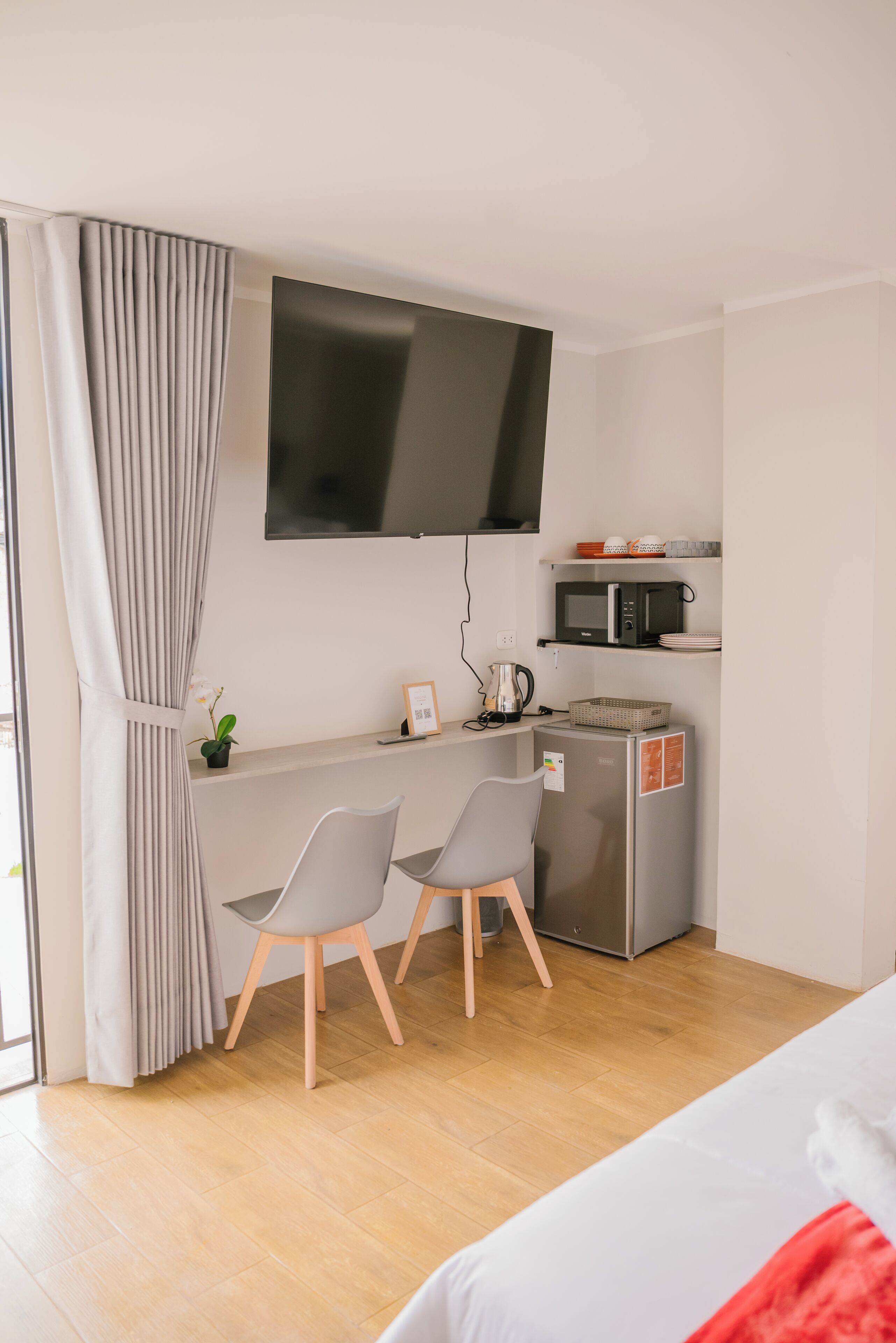 A modern hotel room with a wooden floor, a bed, a desk with two chairs, a flat-screen TV, a microwave, and a small refrigerator.