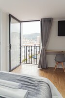 Un dormitorio con cama, escritorio, silla, televisor y balcón con vistas a edificios y montañas.