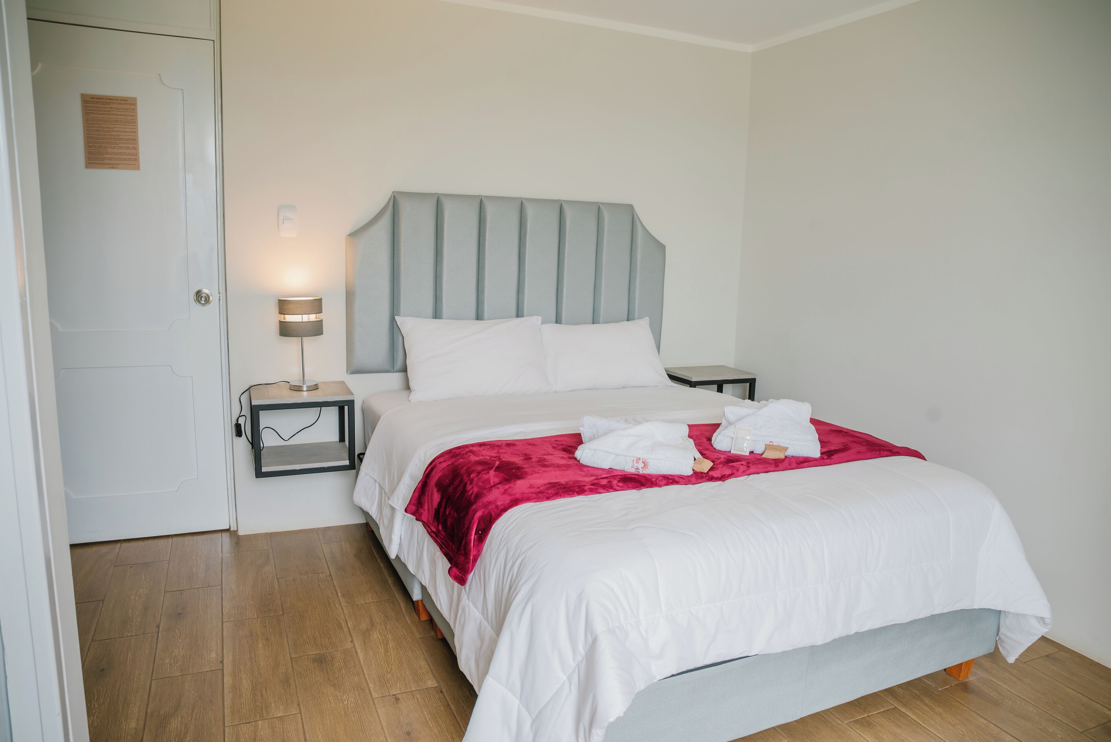 Una cama bien hecha con sábanas blancas y una manta roja, una lámpara de noche y suelo de madera.