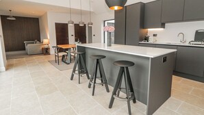 Cozinha moderna com ilha central, banquetas, iluminação pendente e área de jantar com sofá e mesa.