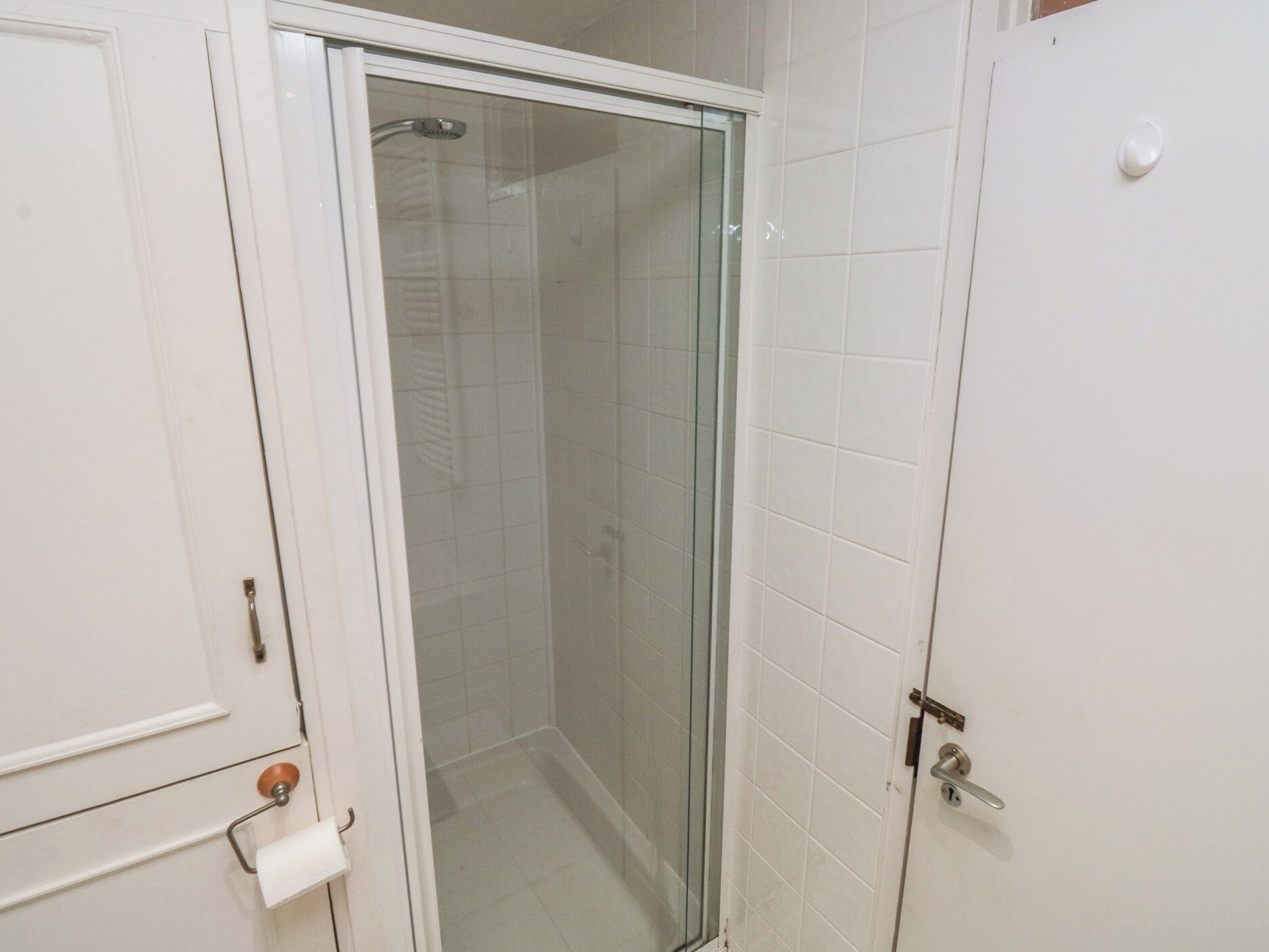 Ein Badezimmer mit Duschkabine aus Glas, weißen Fliesen und einer Tür, an der ein Handtuchhalter und Toilettenpapier angebracht sind.