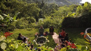 Un groupe de personnes est assis sur une couverture dans un espace extérieur verdoyant et luxuriant, avec une vue sur une étendue d’eau et des collines en arrière-plan.