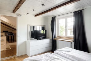 Un dormitorio con cama, televisión, espejo, ventana con cortinas y radiador.