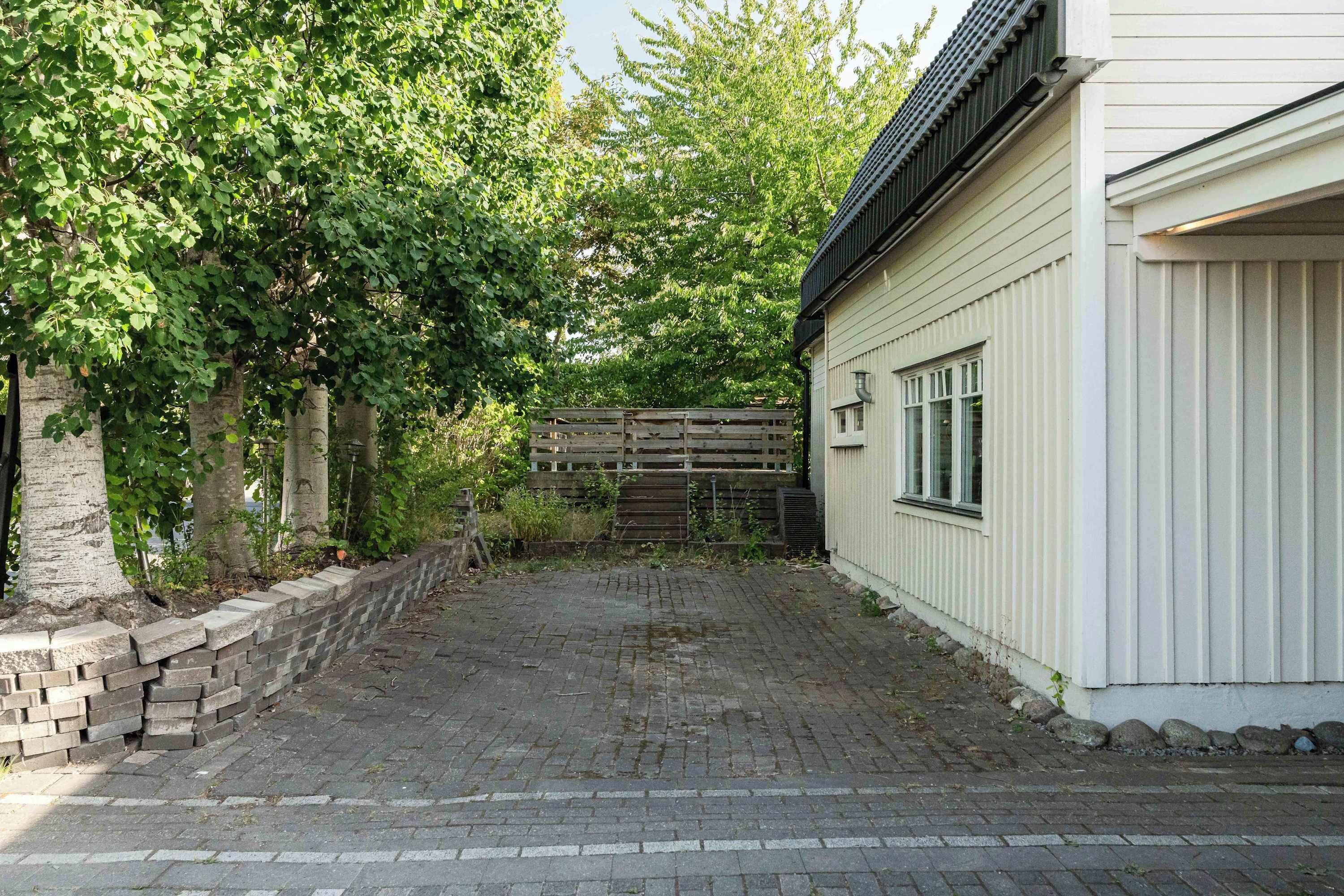 Une cour pavée, bordée d’un mur de briques, d’une clôture en bois et d’un bâtiment dont le revêtement extérieur et les fenêtres sont blancs.