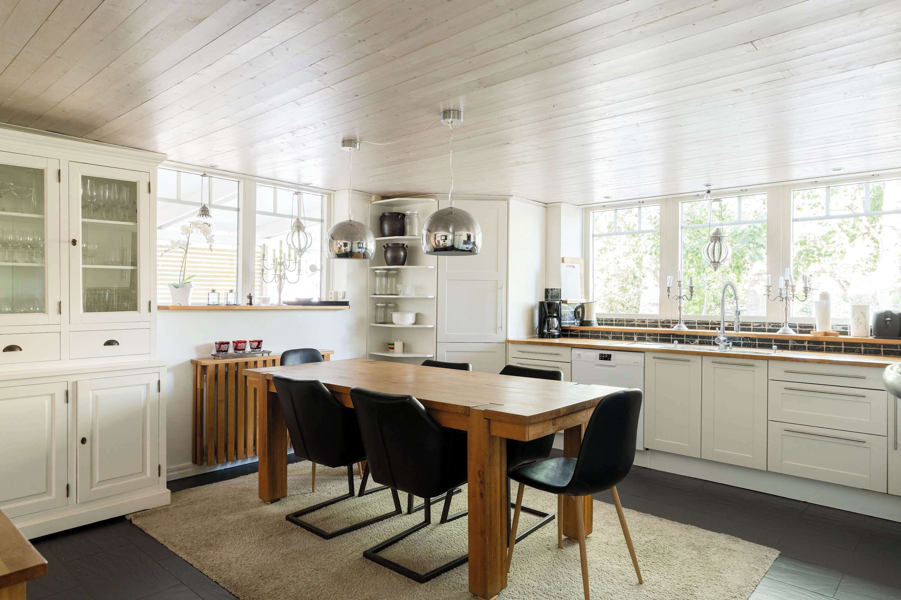 A modern kitchen with white cabinetry, a wooden dining table, black chairs, and a view of greenery outside.