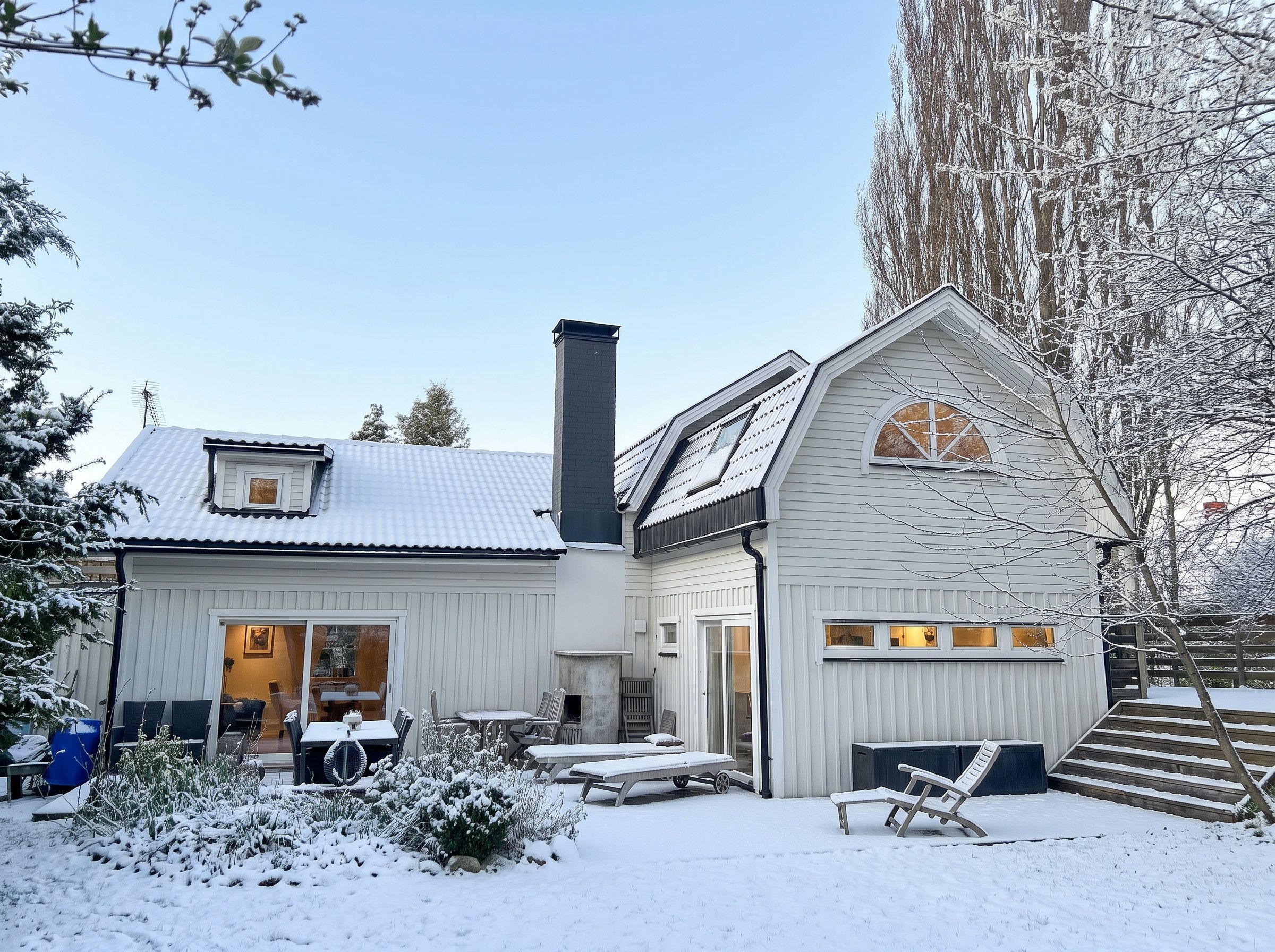 Une maison à deux étages, dont le toit est recouvert de neige, avec une cheminée. On y trouve une terrasse avec des meubles d’extérieur.