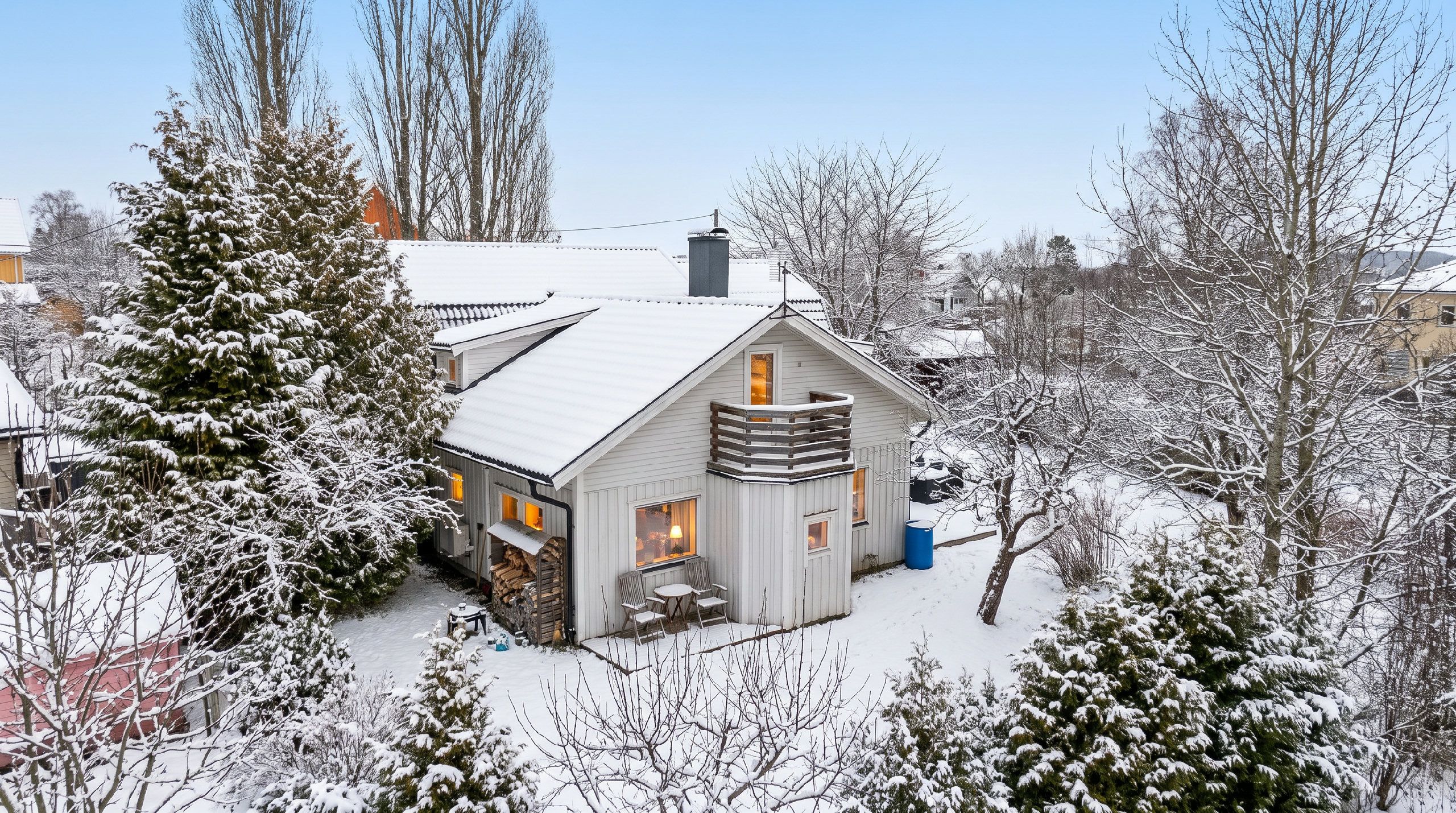 Ett snötäckt hus med en synlig skorsten, en liten veranda med stolar och en hög med ved utanför.