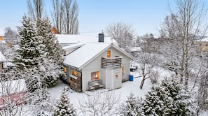 Una casa cubierta de nieve con una chimenea visible, un porche pequeño con sillas y una pila de leña afuera.