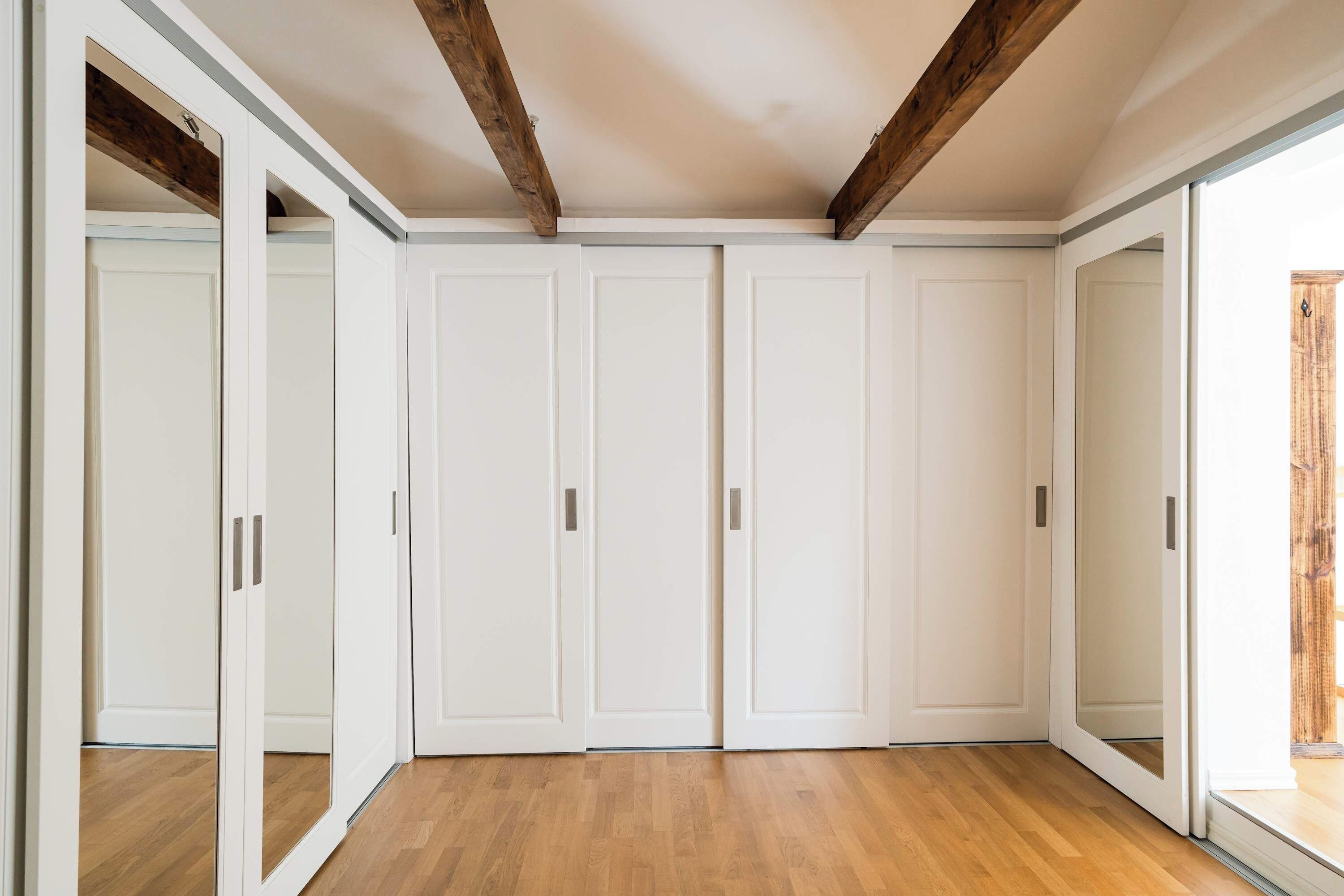 Une pièce dotée d’un plancher en bois, de murs blancs et d’un plafond avec des poutres apparentes. On y trouve trois portes blanches et une porte-fenêtre coulissante.
