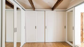 Une pièce dotée d’un plancher en bois, de murs blancs et d’un plafond avec des poutres apparentes. On y trouve trois portes blanches et une porte-fenêtre coulissante.