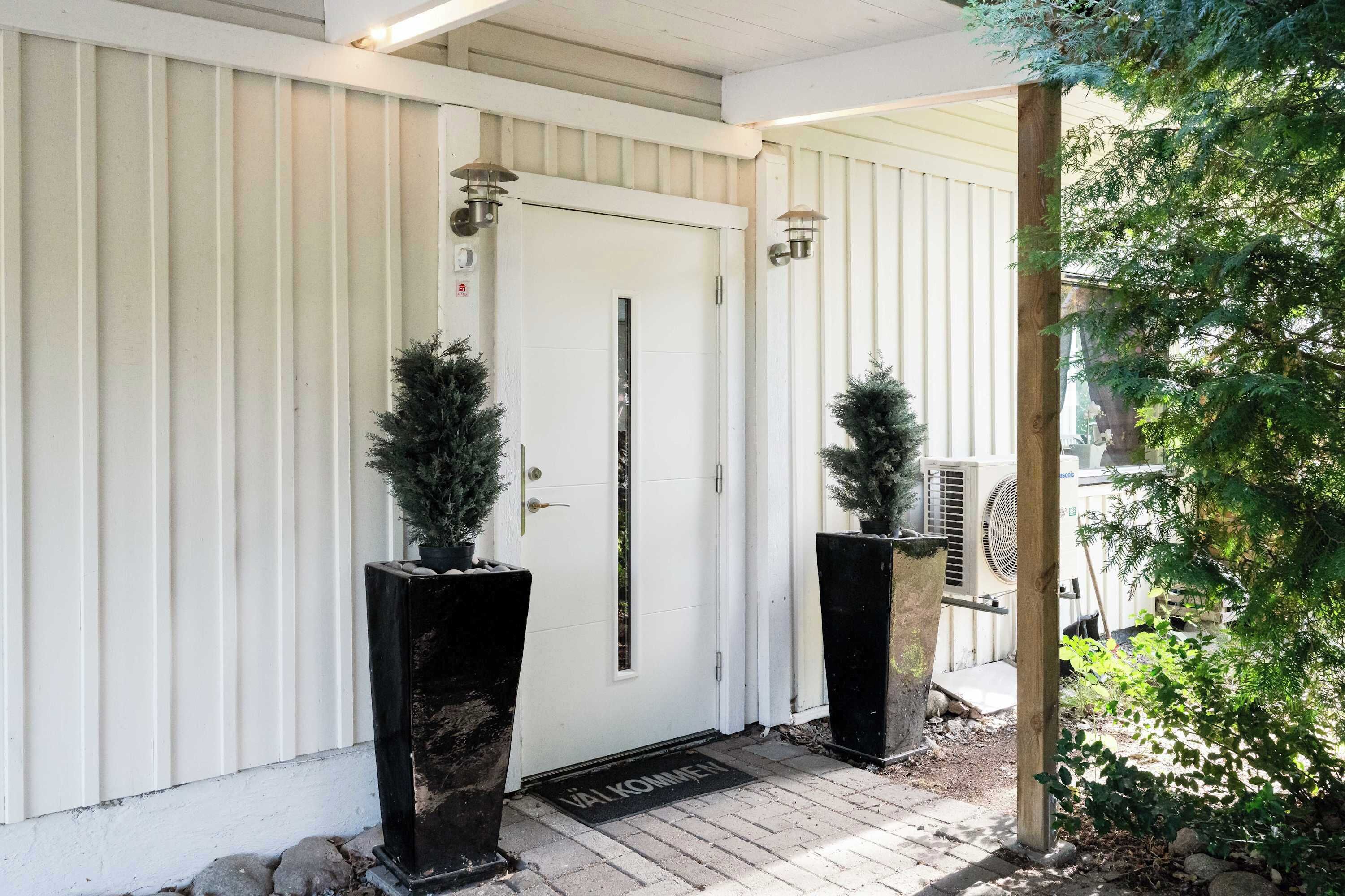 A white door with a vertical stripe, flanked by two black planters with coniferous plants, and a wooden post with a metal bracket.