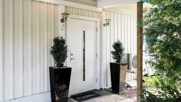 A white door with a vertical stripe, flanked by two black planters with coniferous plants, and a wooden post with a metal bracket.