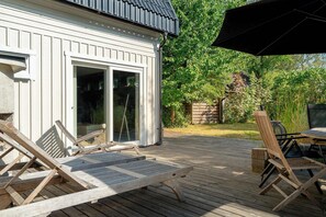 A deck with lounge chairs and a table, a white building with a sliding glass door, and a garden with trees and a wooden fence.