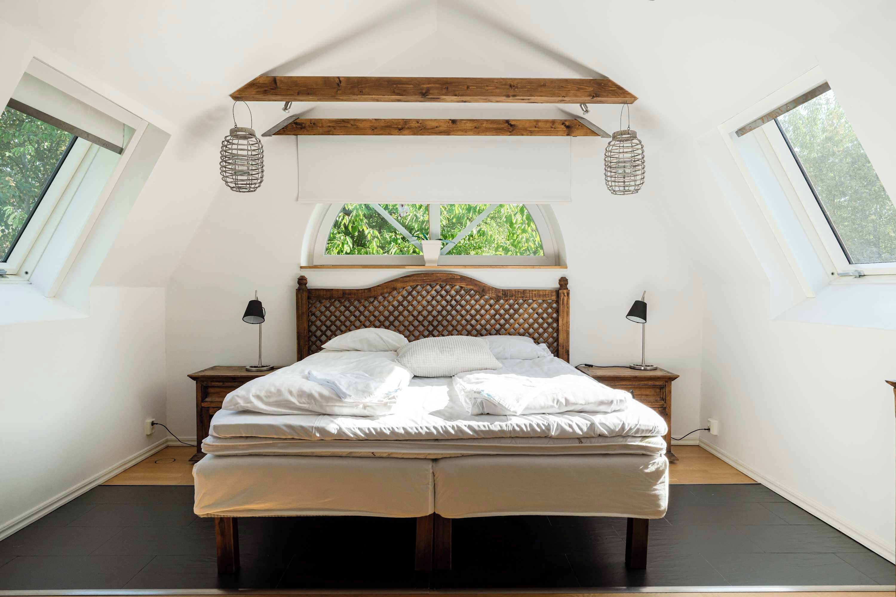 Een slaapkamer met een schuine kap, een houten hoofdbord, een bed met witte lakens, twee nachtkastjes met lampen en een raam met uitzicht op groen.