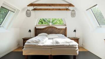 Een slaapkamer met een schuine kap, een houten hoofdbord, een bed met witte lakens, twee nachtkastjes met lampen en een raam met uitzicht op groen.