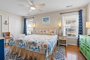 A bedroom with a bed, a chair, a nightstand, a dresser, a ceiling fan, and a window with curtains. - Salty Venture 𓇼 Game Room + Fire Pit + Family Fun (Oak Island)
