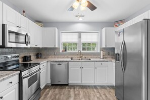 A modern kitchen with white cabinets, stainless steel appliances, a granite countertop, and a window with blinds. - Salty Venture 𓇼 Game Room + Fire Pit + Family Fun (Oak Island)