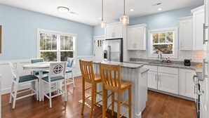 A modern kitchen with white cabinets, a granite countertop, and stainless steel appliances. There is a dining area with a table and chairs, and a large window with blinds. - Sea La Vie𓇼Central Location𓇼Cozy𓇼Fire Pit (Oak Island)
