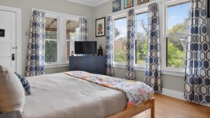 A bedroom with a bed, dresser, TV, and large windows with patterned curtains. - New! Stunning Pet-Friendly Home + 3 Min to DT! (Seguin)