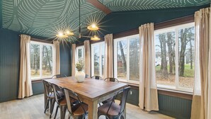 En spisestue med et træbord, seks stole, en vase med blomster og store vinduer med gardiner.