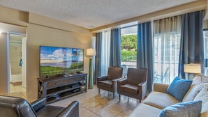 A hotel living room with a flat-screen TV, a sofa, two armchairs, a coffee table, and a view of greenery through large windows. - 3bdrm 3bath Luxury Condo Across from Kamaole Beach Kihei Maui Full AC Sleeps 8 (Kihei)