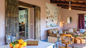 A rustic interior with a wooden door, a dining table with oranges and a chessboard, a sofa with cushions, and a large floral artwork on the wall.