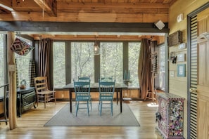 L’intérieur chaleureux d’un chalet avec une table à manger en bois, deux chaises bleues, une cheminée et de grandes fenêtres donnant sur la forêt.