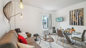Una sala de estar moderna con un sofá, una mesa de comedor con sillas, un televisor montado en la pared y una ventana con cortinas.