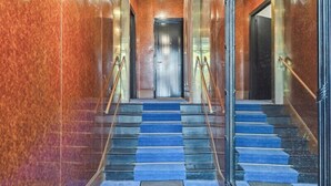 Una imponente escalera con alfombra azul que conduce a una puerta, rodeada de paredes de mármol pulido y un espejo grande.