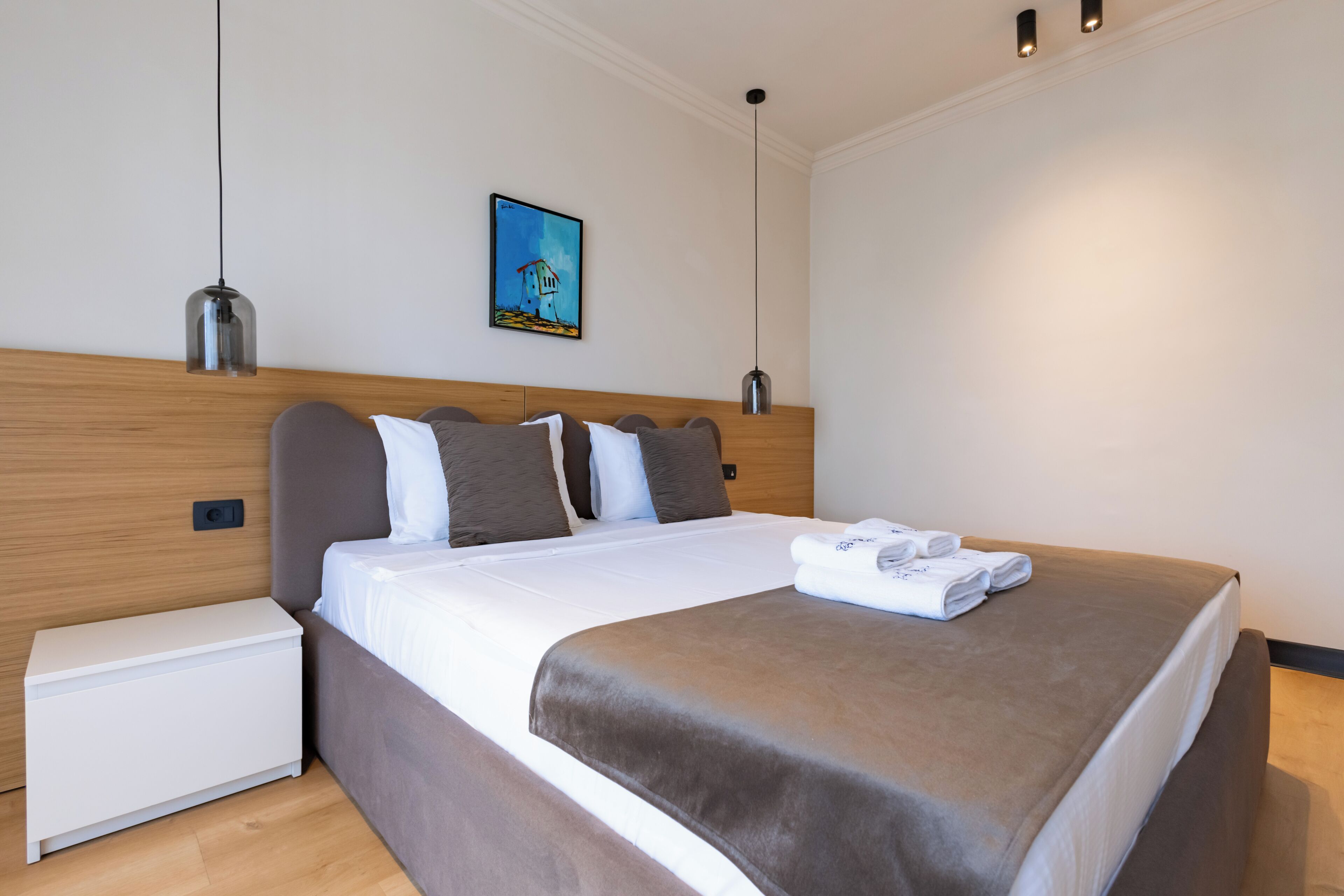 A modern hotel room with a wooden headboard, a neatly made bed, a bedside table, and a wall-mounted painting.
