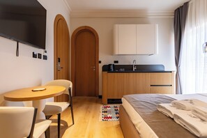 A hotel room with a bed, a desk, a chair, a TV, a kitchenette, and a bathroom door. - PRI ApartHotel (Pristina)
