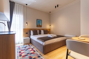 A hotel room with a bed, bedside tables, a desk, a chair, a heater, and a painting on the wall. - PRI ApartHotel (Pristina)